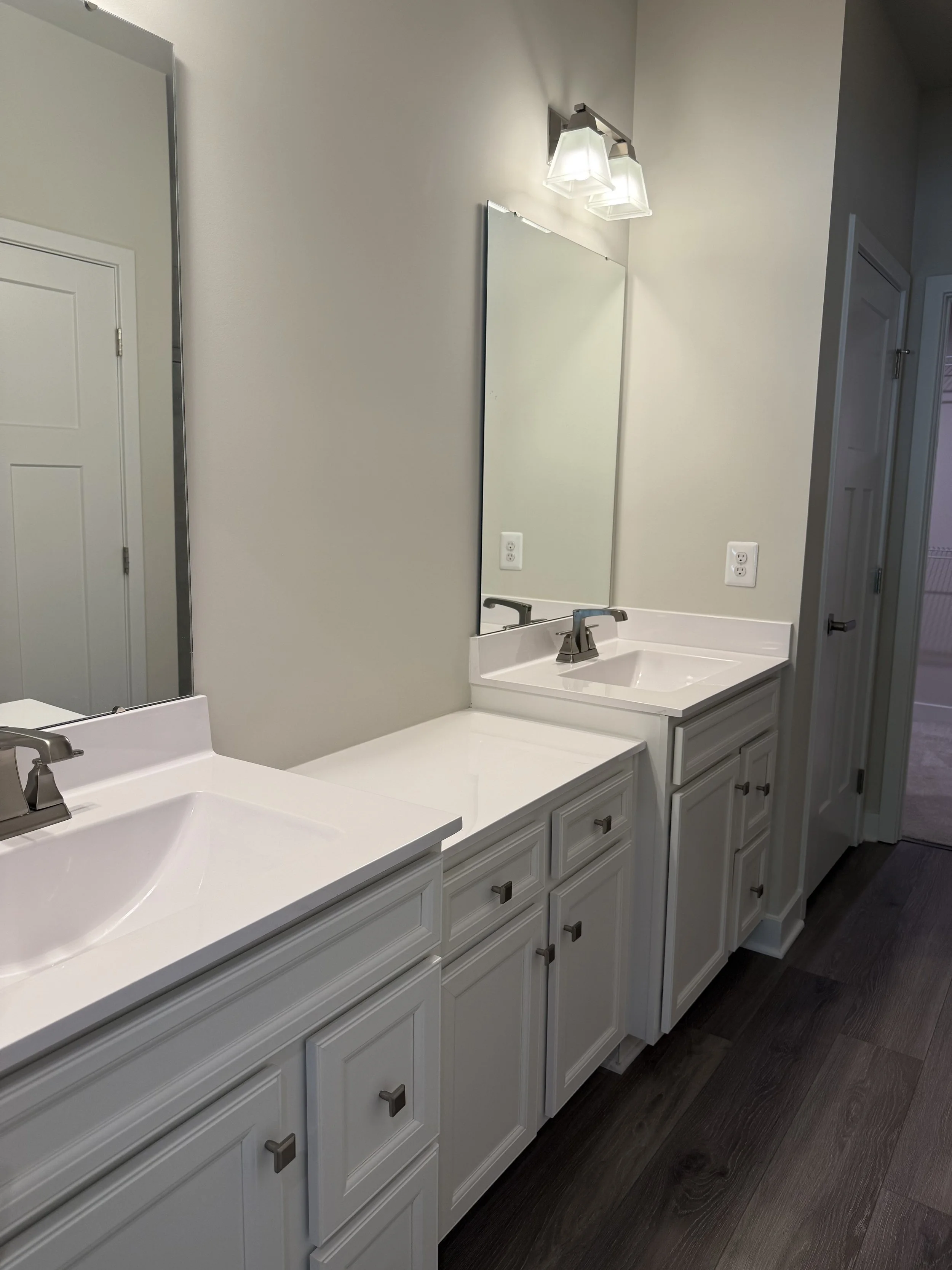 Bathroom vanity with multiple white cabinets, two sinks, and a large mirror, illuminated by three lights, with a dark wood floor.