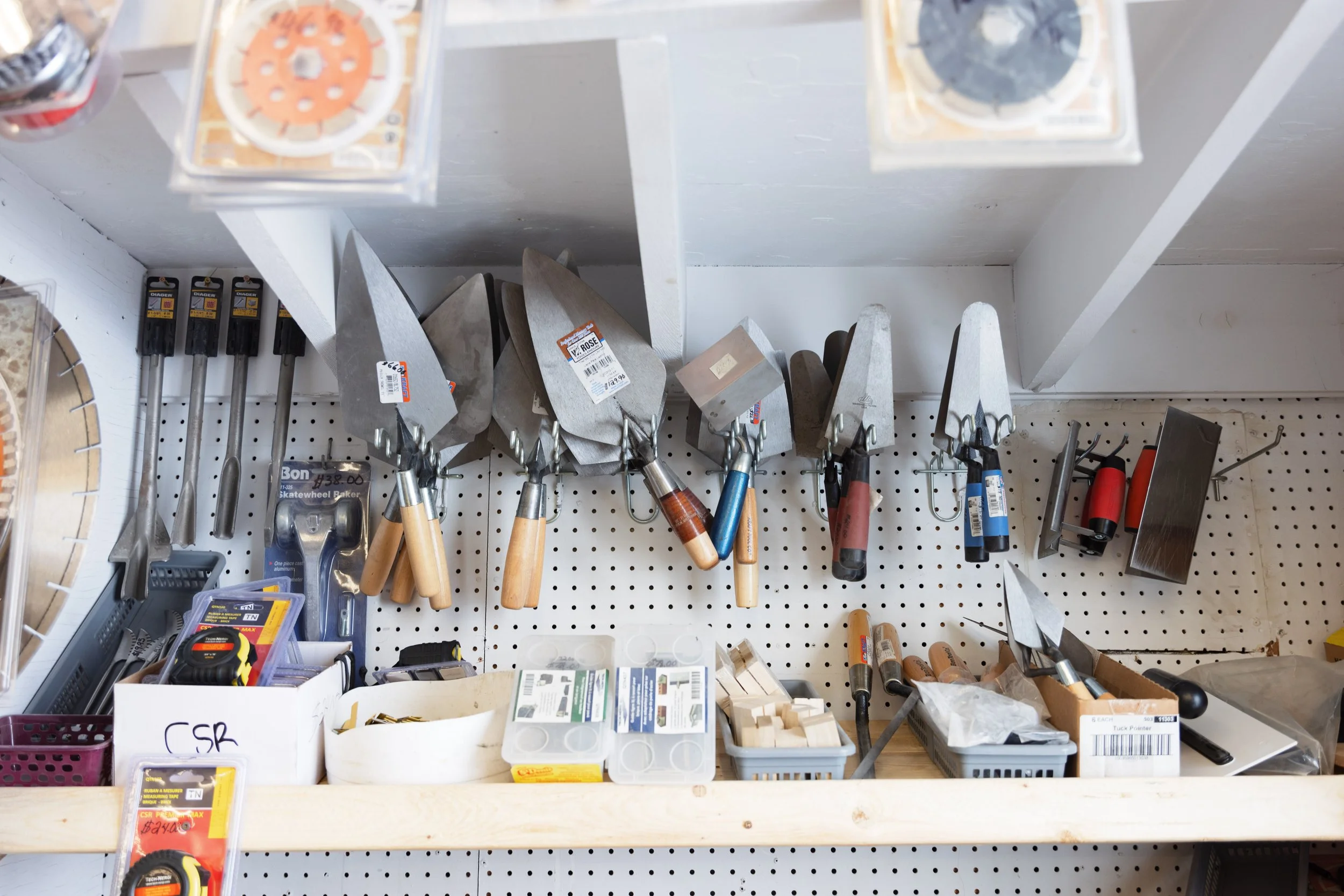 A workshop pegboard holding gardening tools such as trowels, cultivators, and other hand tools, with other small storage containers and boxes on the workbench below.