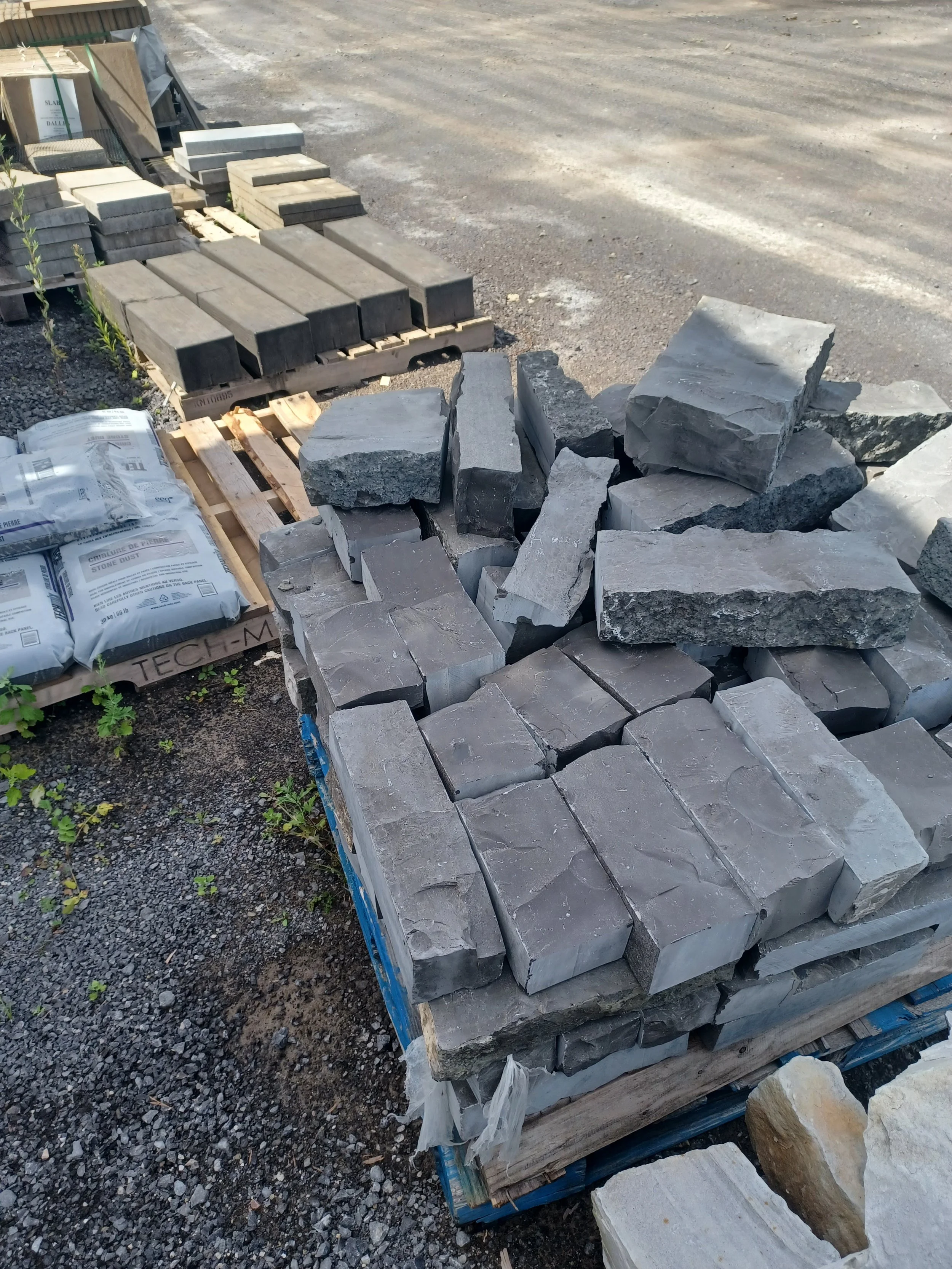 Stacks of building and paving stones, including gray stones in various shapes and sizes, arranged on pallets outdoors, with additional materials and gravel nearby.