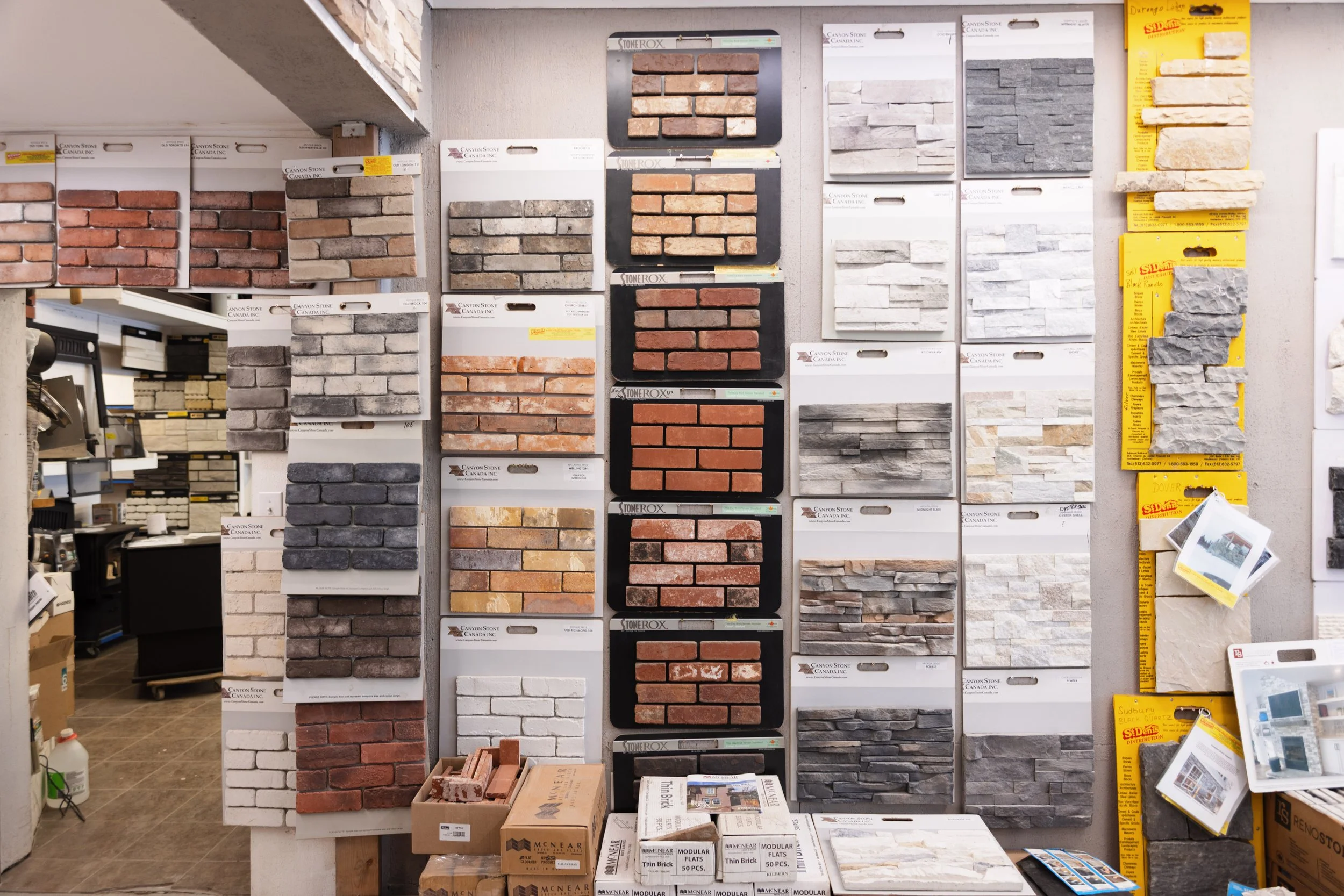 Display of various brick and stone veneer samples on a wall, with samples arranged in rows of different colors and textures in a store. Shelves with boxes of additional building materials are below.