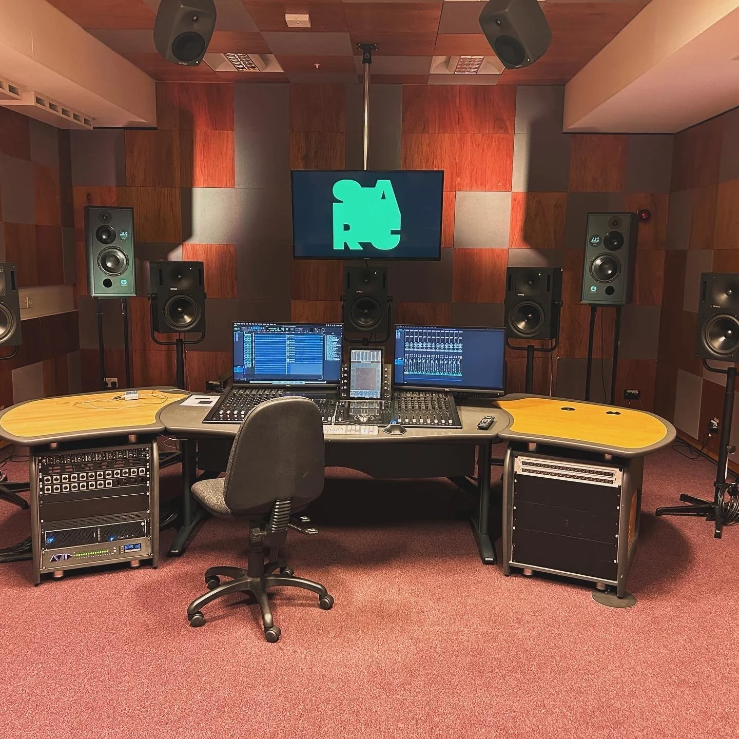 A professional music recording and mixing studio with a large mixing console, two computer screens displaying audio software, multiple studio monitors, and acoustic paneling on the walls.