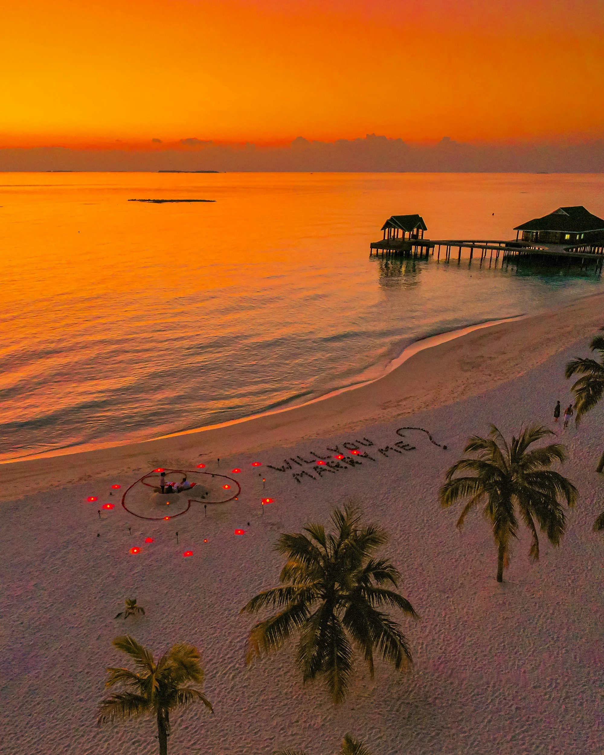 A beach at sunset with palm trees, a pier with a structure, and candles arranged on the sand spelling out 'Will You Marry Me?'.