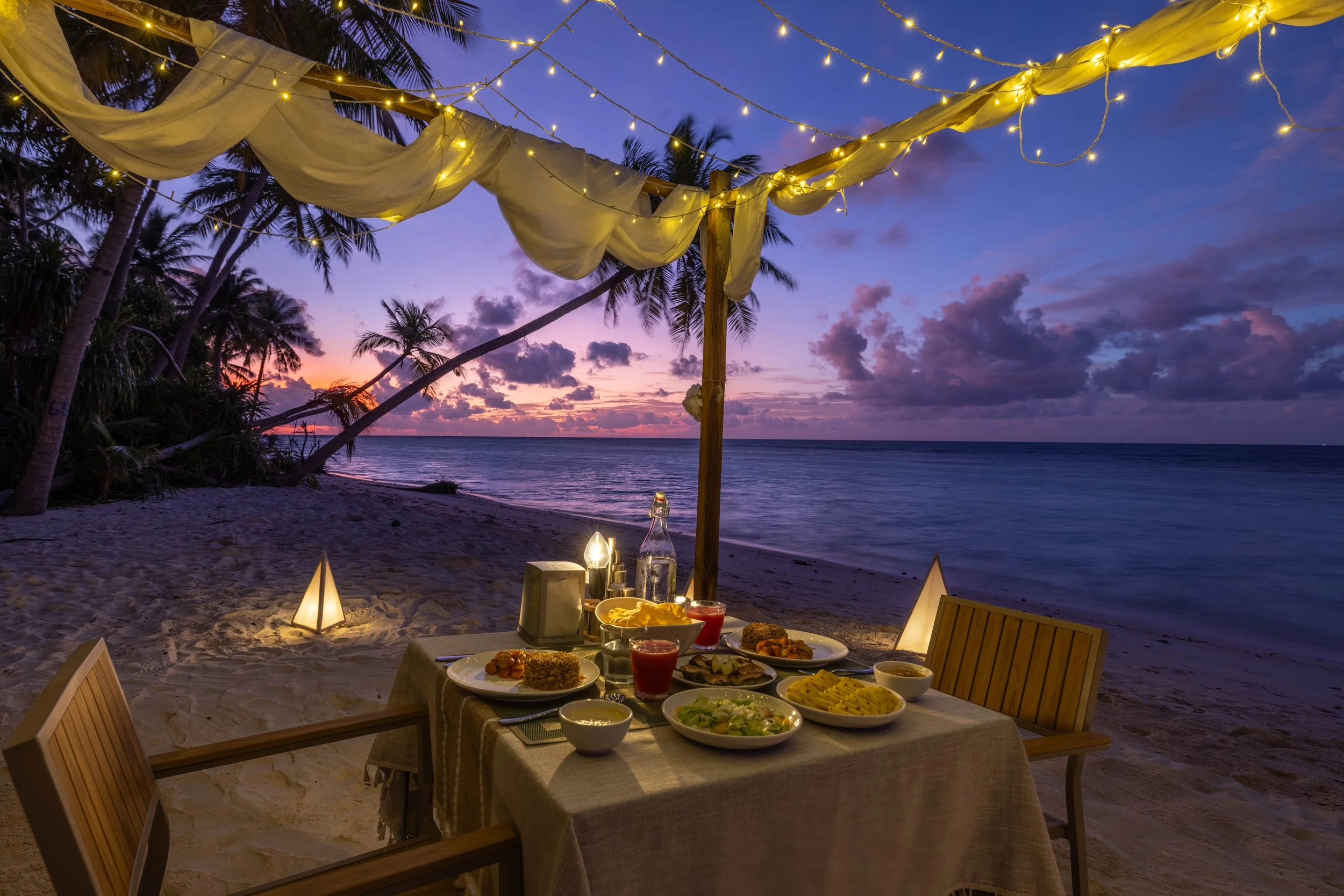 A dining table set with plates of food, drinks, and candles on a beach at sunset. Overhead string lights and a canopy are visible, with palm trees and the ocean in the background.