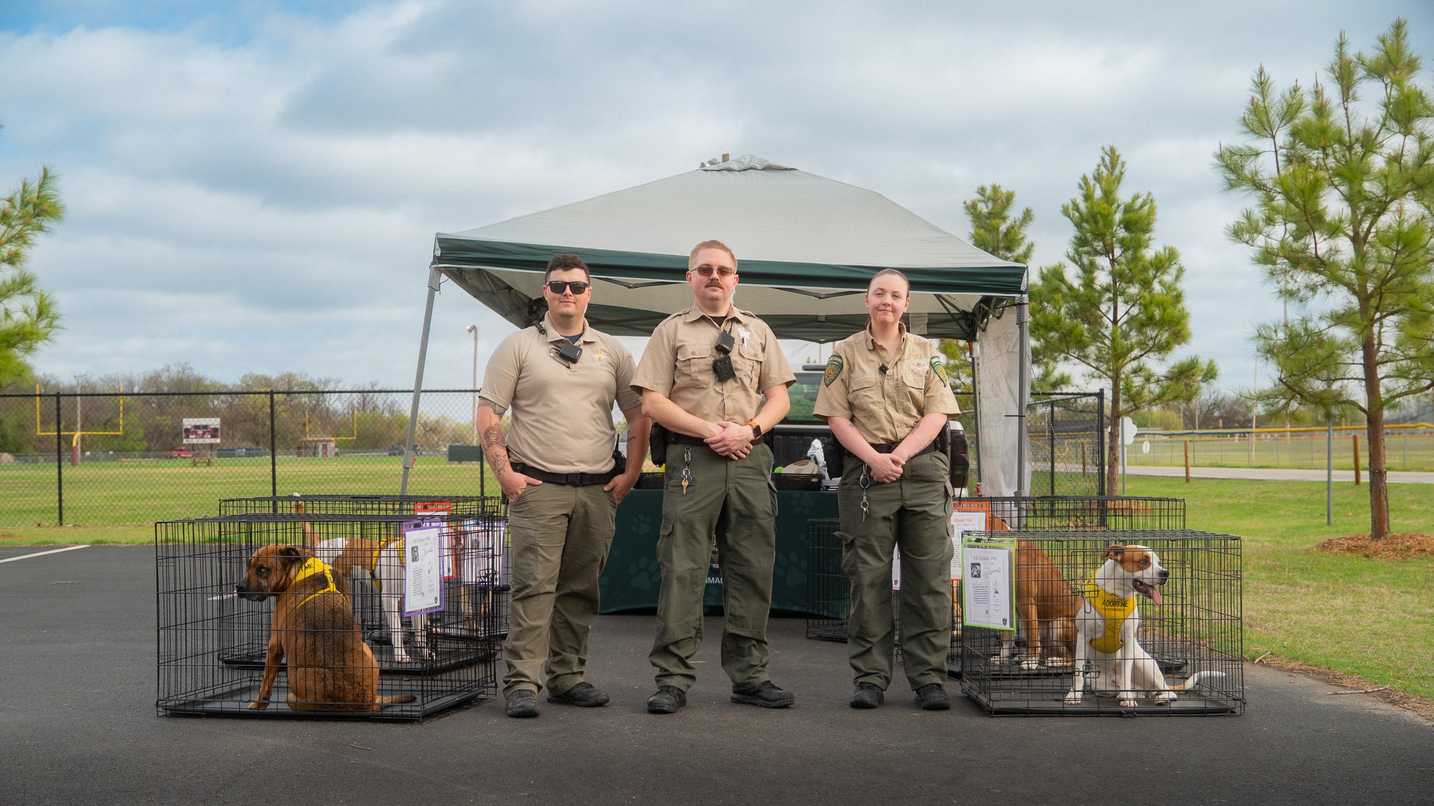 Three wildlife officers standing in front of a tent at an outdoor event, with three dogs in cages labeled 'Adopt Me' in a grassy park area, trees, and cloudy sky in the background.