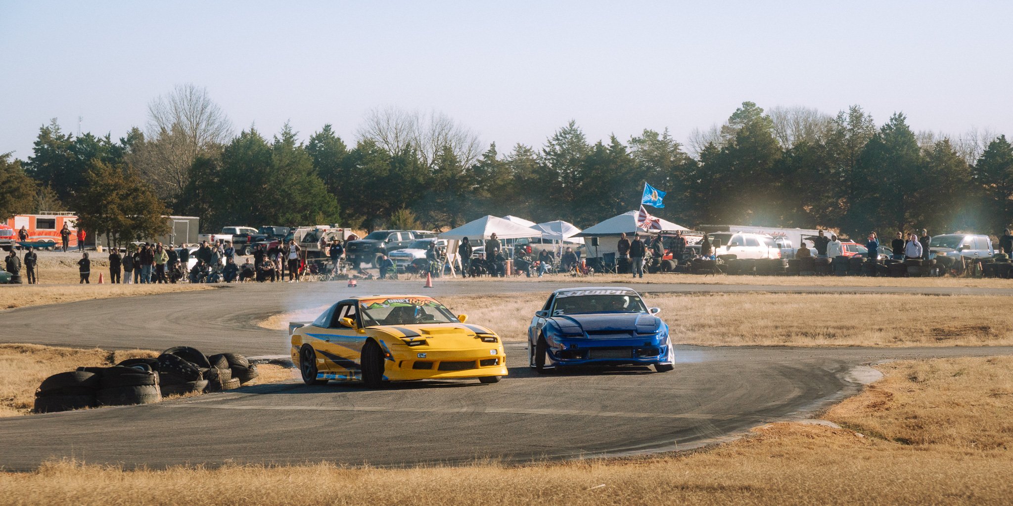 Two drift cars, one yellow and one blue, drifting on a racetrack during a racing event. Spectators and tents are visible in the background, with a crowd watching the race.