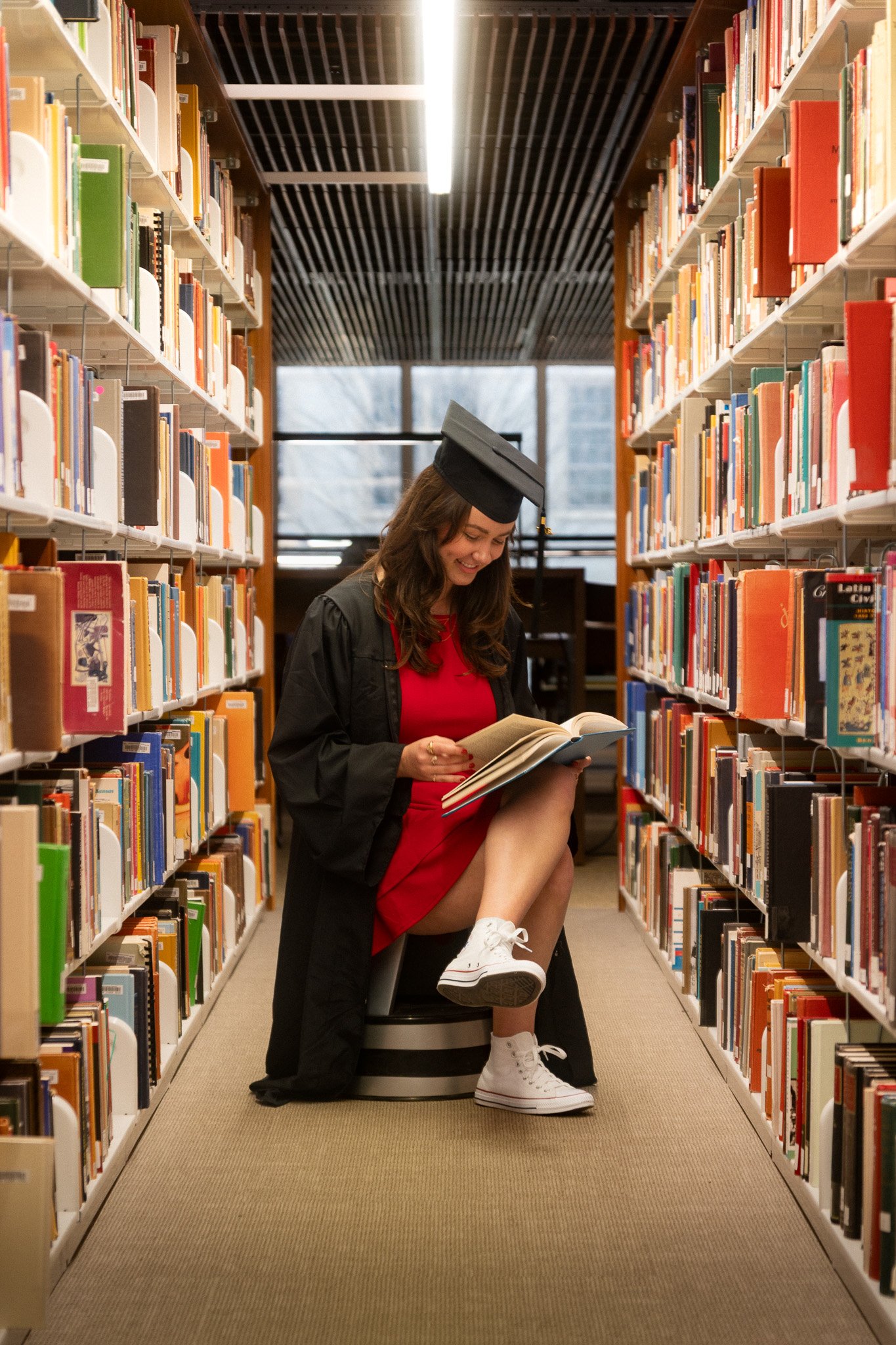 A woman in a graduation cap and gown is smiling while reading a book in a library. She has one leg raised and resting on a chair, wearing a red dress and white sneakers. The library has tall shelves filled with books, and a window in the background.