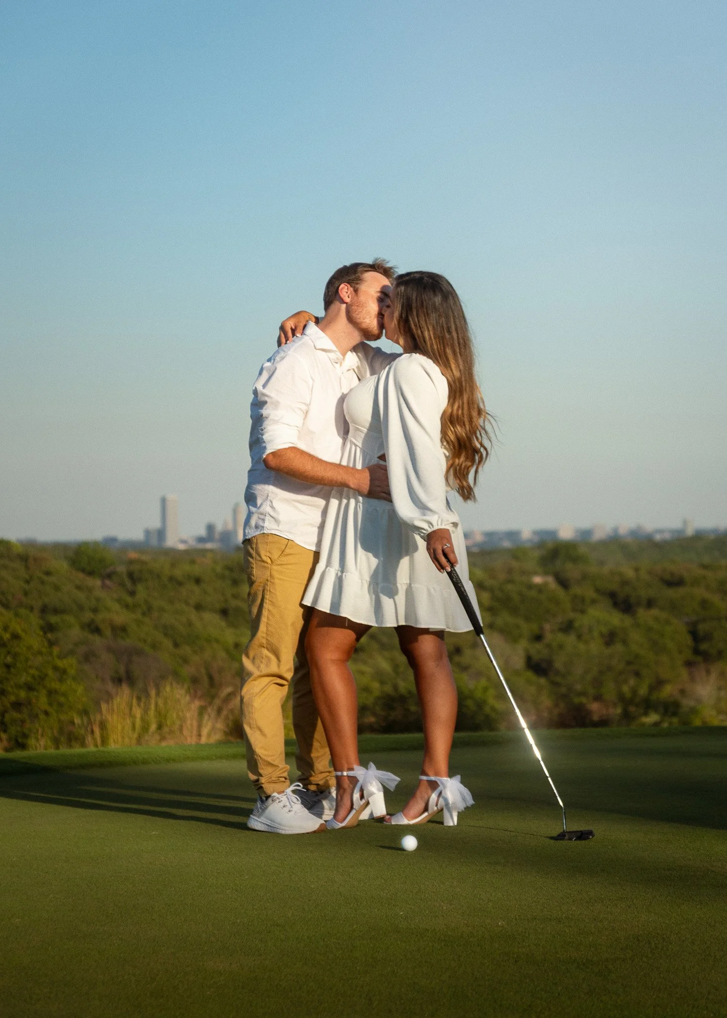 A couple kissing on a golf course before putting with a golf club in hand. The woman is wearing white high heels with bows, and the man is wearing white sneakers. The background shows a city skyline and clear sky.