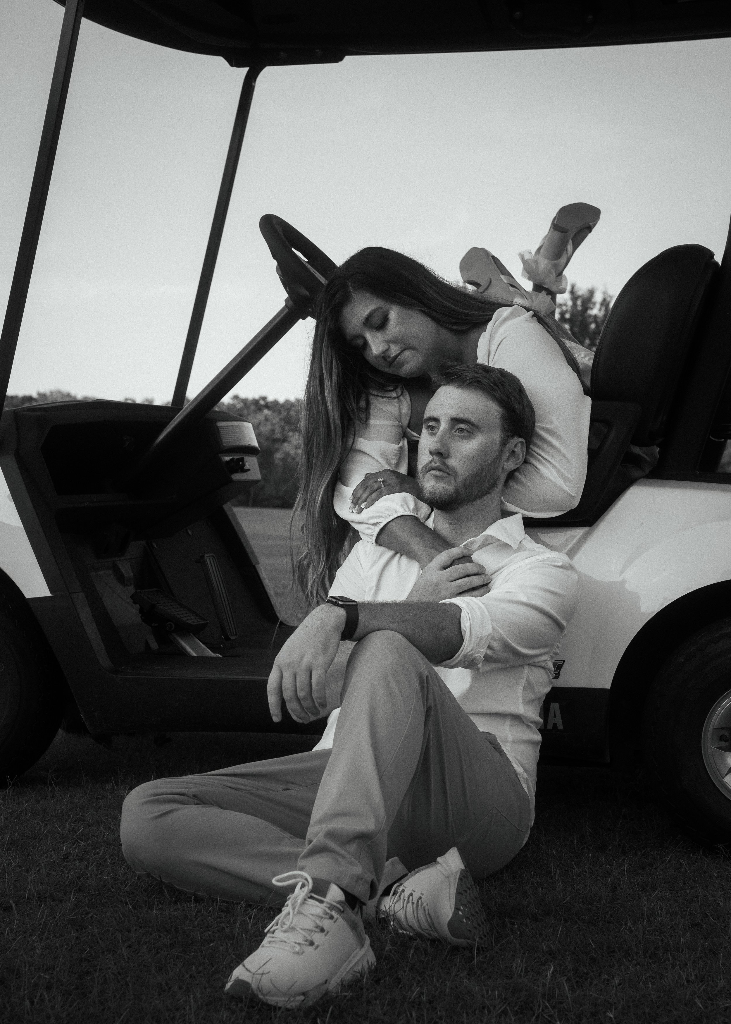 Black and white photo of a young man sitting on the grass with his back against a golf cart, and a young woman leaning over him, inside the golf cart. The woman is resting her head on the man's shoulder while he looks off into the distance. The woman has long hair and is wearing a light-colored jacket, while the man has short hair, a beard, and is wearing a light-colored shirt and pants. The photo has a calm and intimate atmosphere.