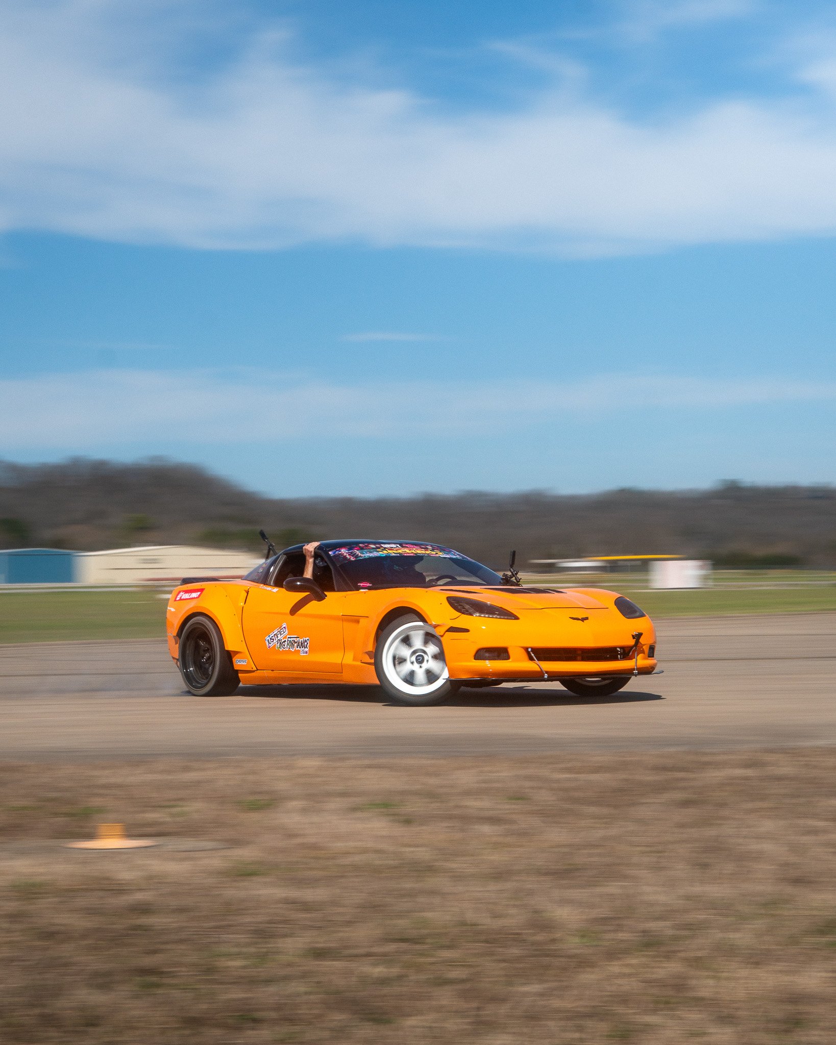A bright orange Corvette drifting on a track with a background of mountains, sky, and clouds.