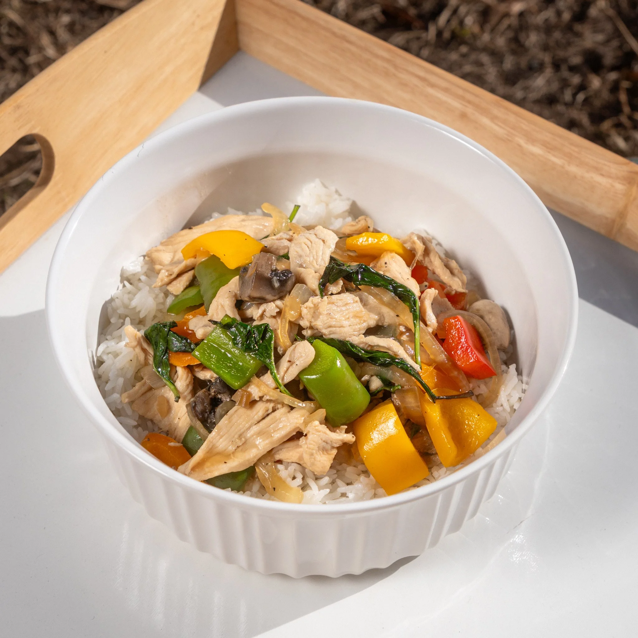 A white cermaic bowl filled with cooked white rice topped with cooked vegetables and shredded chicken, placed on a white surface with a wooden tray in the background.