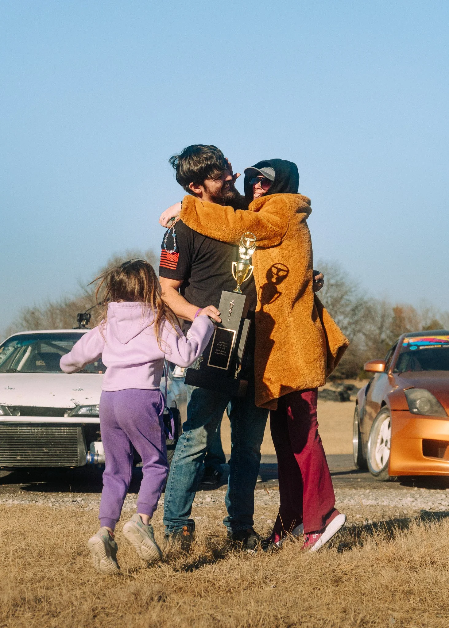 A couple hugging, one wearing a brown fuzzy jacket and sunglasses, the other with curly hair and a beard, holding a trophy. A young girl in purple clothes is standing nearby, and there are parked cars in the background outdoors on a grassy field.