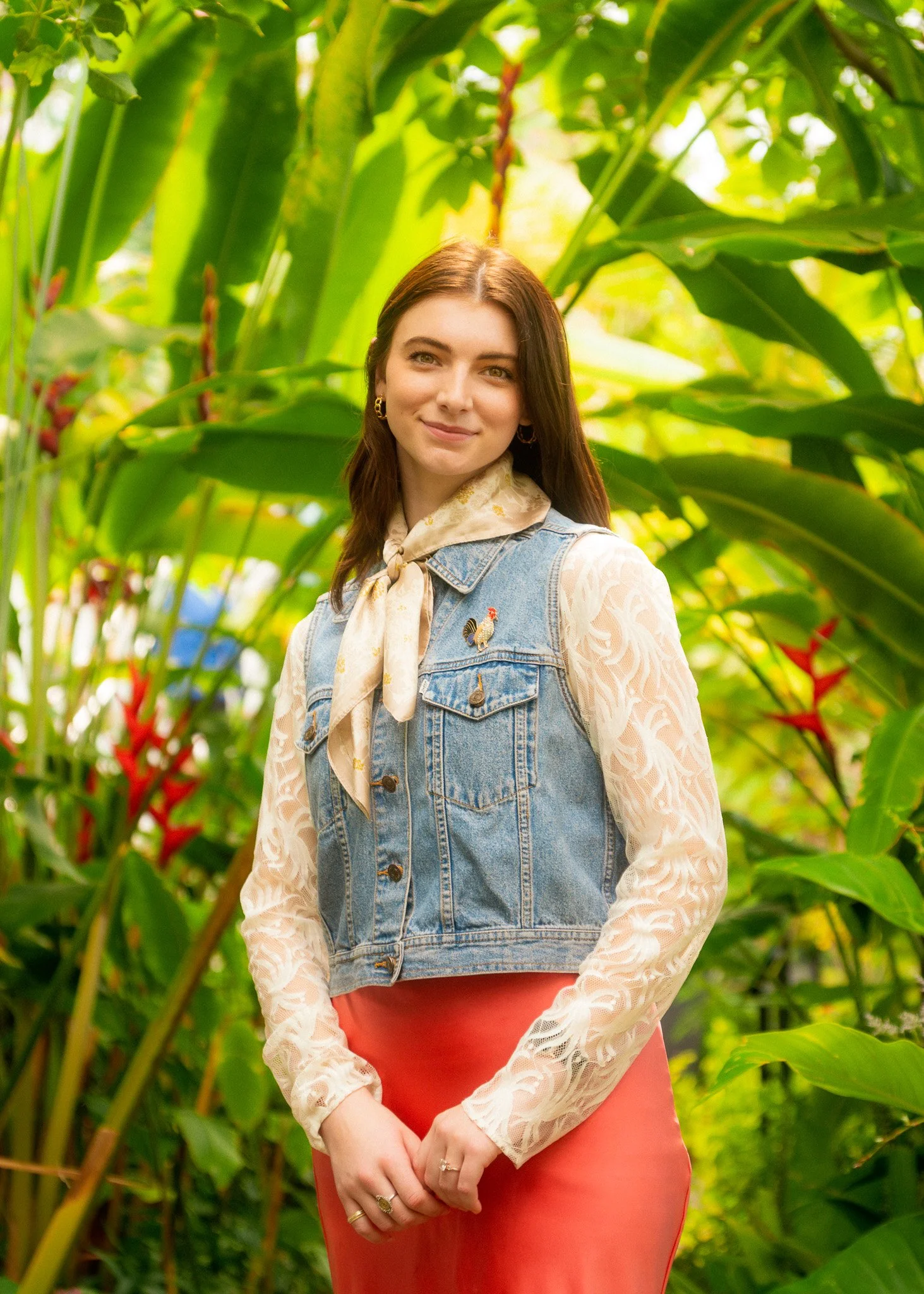 A young woman standing in a lush green jungle, wearing a denim vest over a sheer, long-sleeved top, a light scarf tied around her neck, and a coral-colored skirt. She has brown hair, subtle makeup, and is smiling.