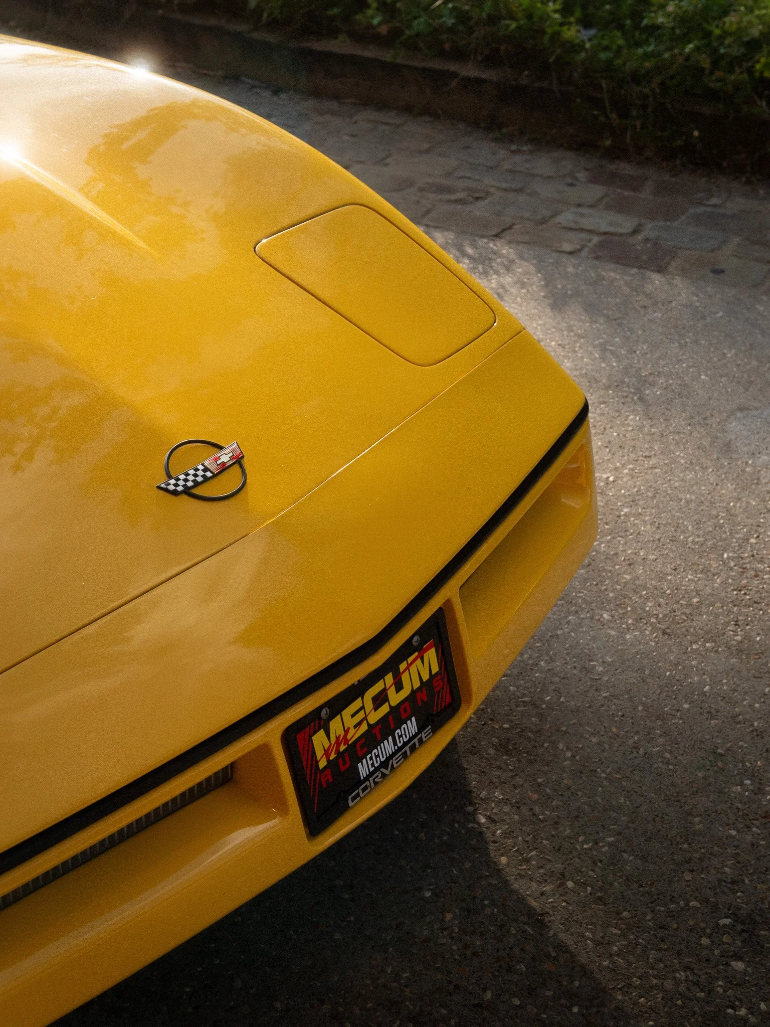 Close-up of a yellow Corvette car hood with a Corvette emblem and a license plate with promotional text for Mecum auctions.