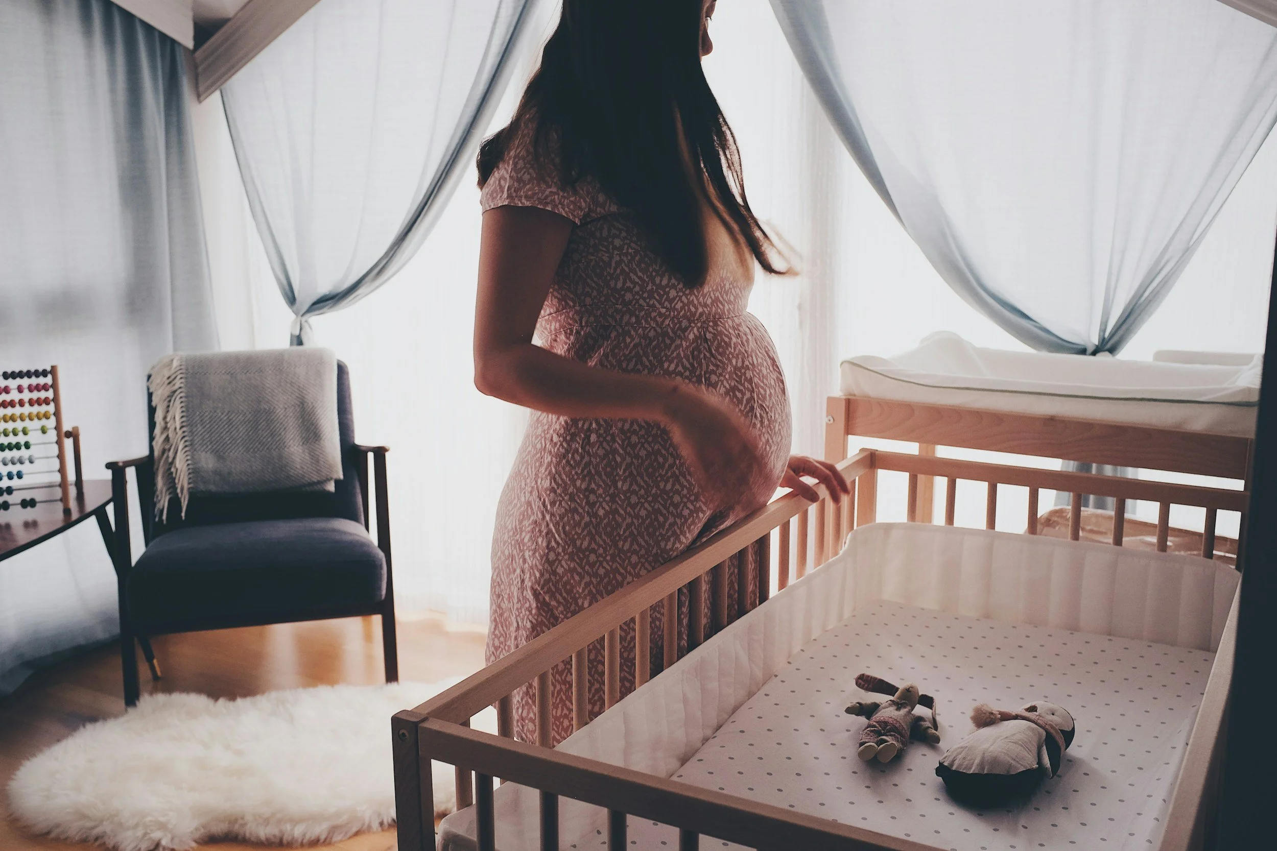 A pregnant woman standing next to a crib in a nursery with white curtains, a chair, and a fluffy white rug.