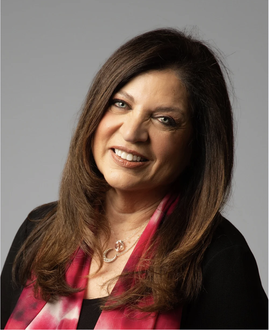 Portrait of a woman with long brown hair, wearing a black top and pink scarf, smiling at the camera against a gray background.