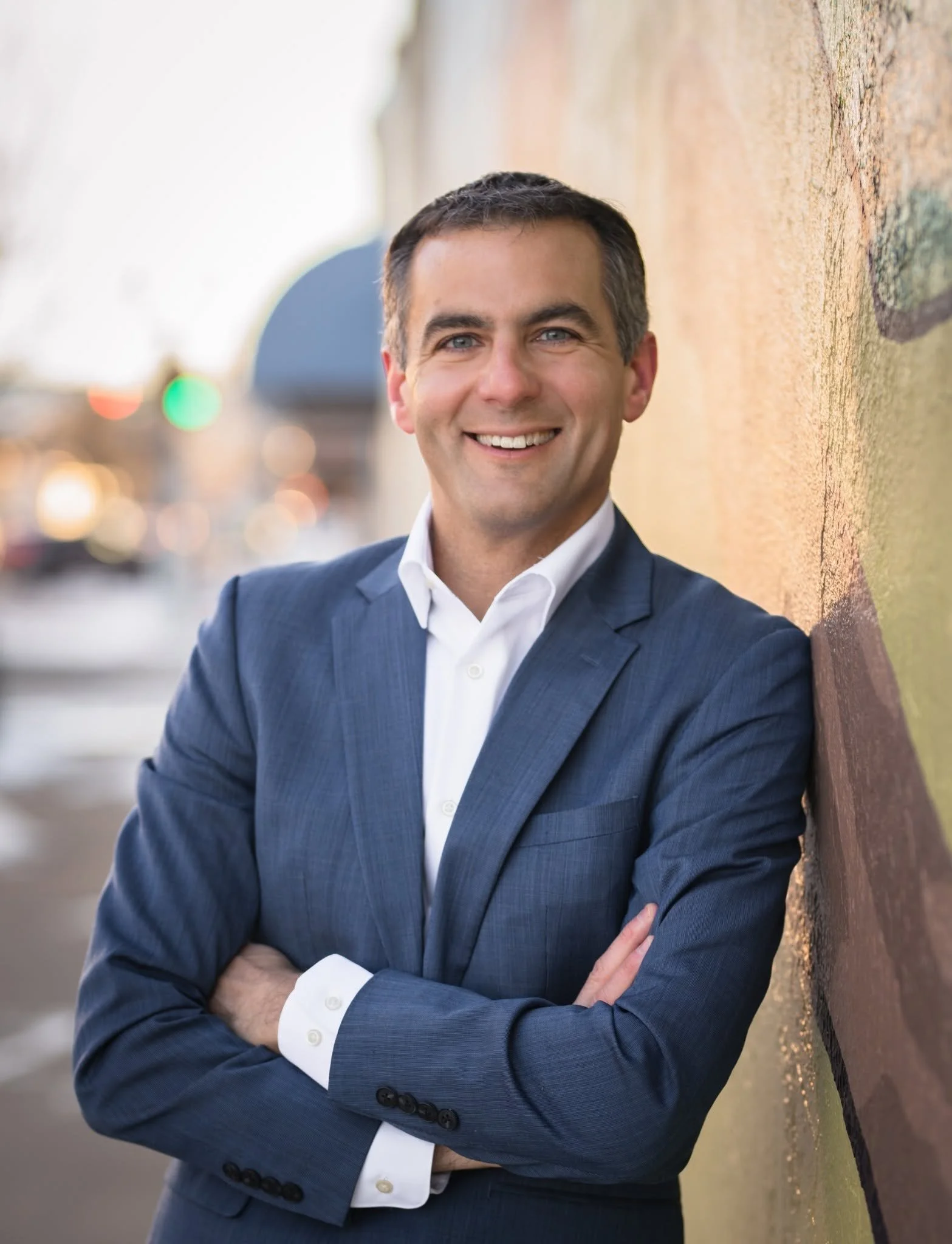A man in a blue suit with a white shirt standing with his arms crossed, leaning against a textured brick wall outdoors during daytime, smiling and looking directly at the camera.