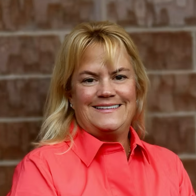 Portrait of a smiling woman with blonde hair wearing a red blouse against a brick wall background.