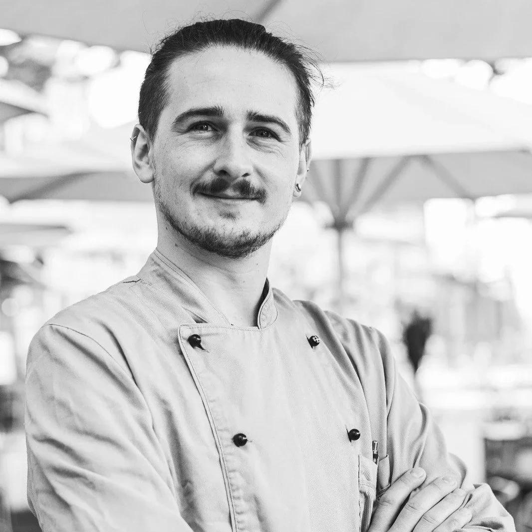Schwarz-weiß-Foto eines jungen männlichen Küchenchefs, der lächelnd mit verschränkten Armen vor einem gastronomischen Hintergrund steht.