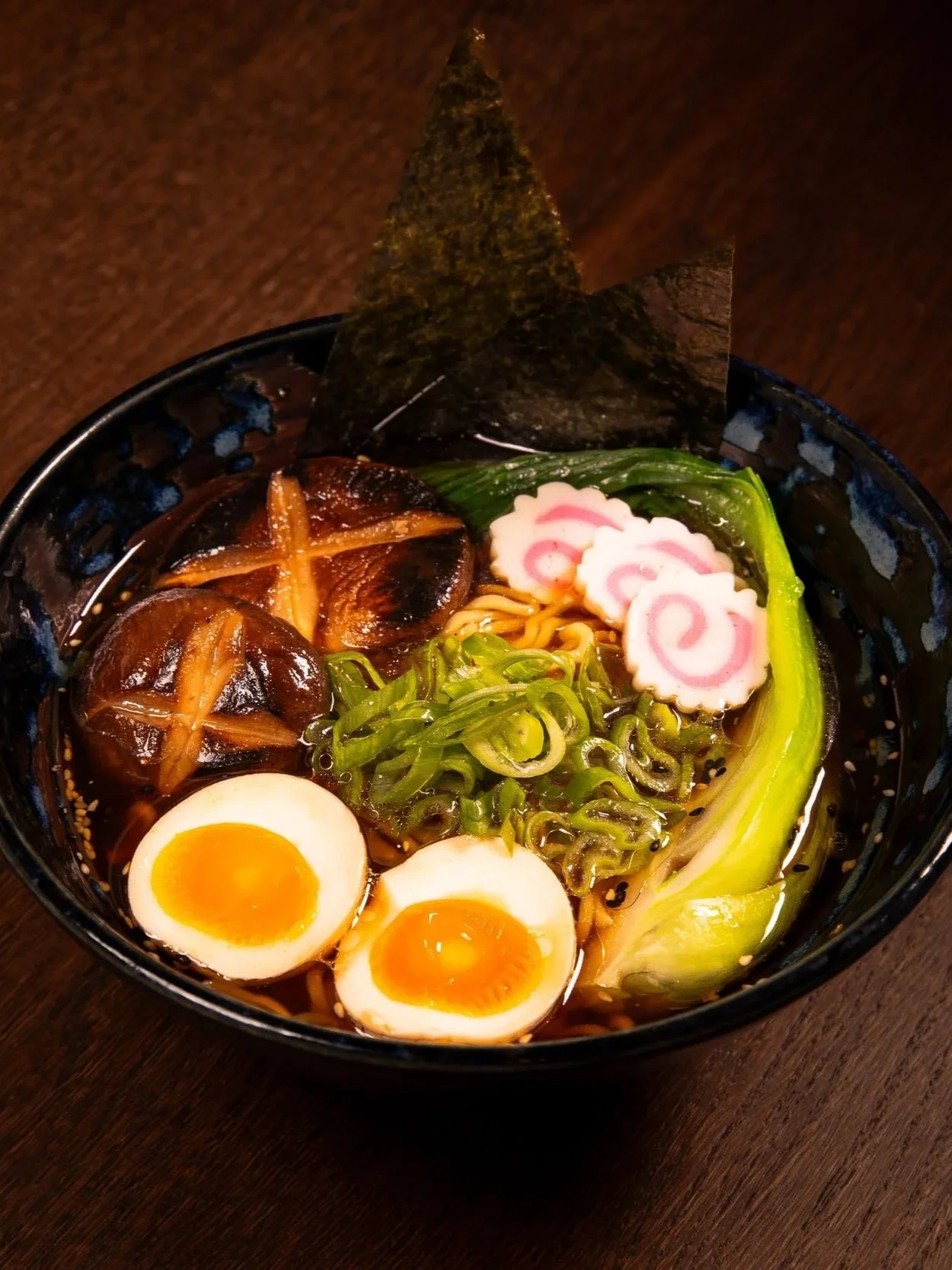 Schüssel mit Ramen-Nudeln, halbierte weichgekochte Eier, gegrillte Shiitake-Pilze, Frühlingszwiebeln, Nori-Algen, Kamaboko (japanischer Fischkuchen) und gekochtem Lauch.
