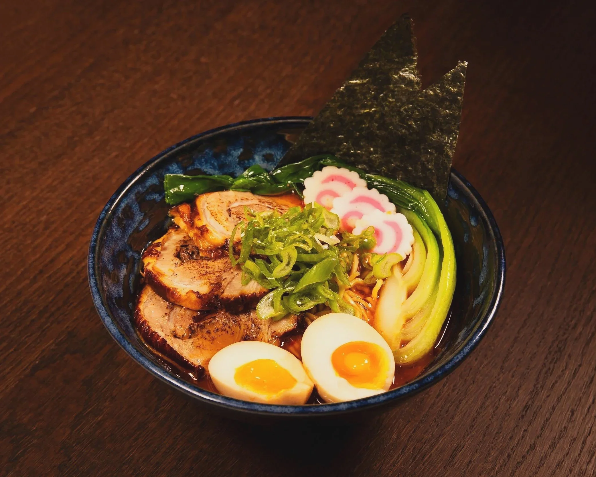 Schüssel mit Ramen-Nudeln, gekochtem Ei, Frühlingszwiebeln, Rindfleisch, Nori-Blatt, Kamaboko und Gemüse auf einem dunklen Holztisch.