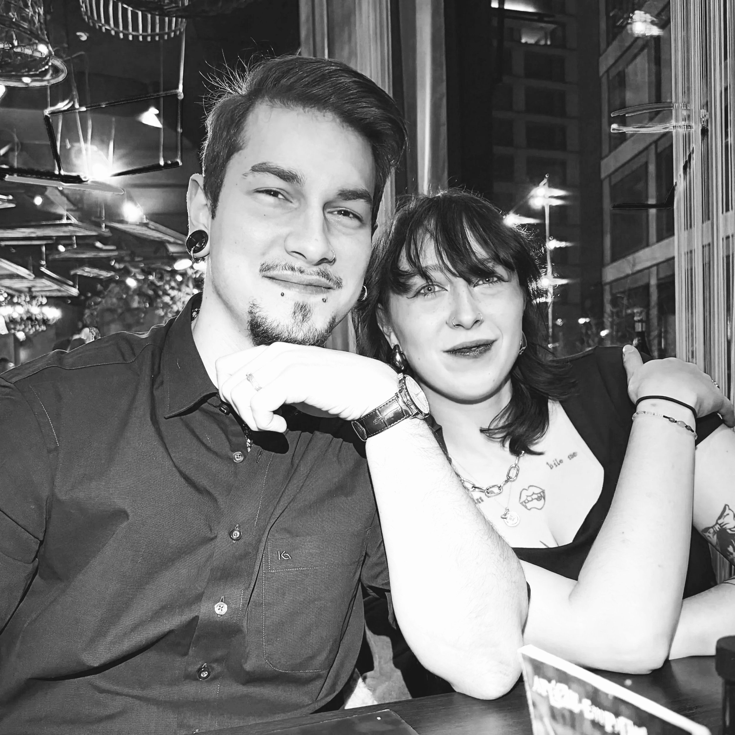 Zwei Menschen, eine mit kurzen dunklen Haaren und Piercings am Gesicht, eine mit schwarzen kurzen Haaren und einem Halsband, sitzen in einem Restaurant oder Café, während sie miteinander sprechen und lachen.