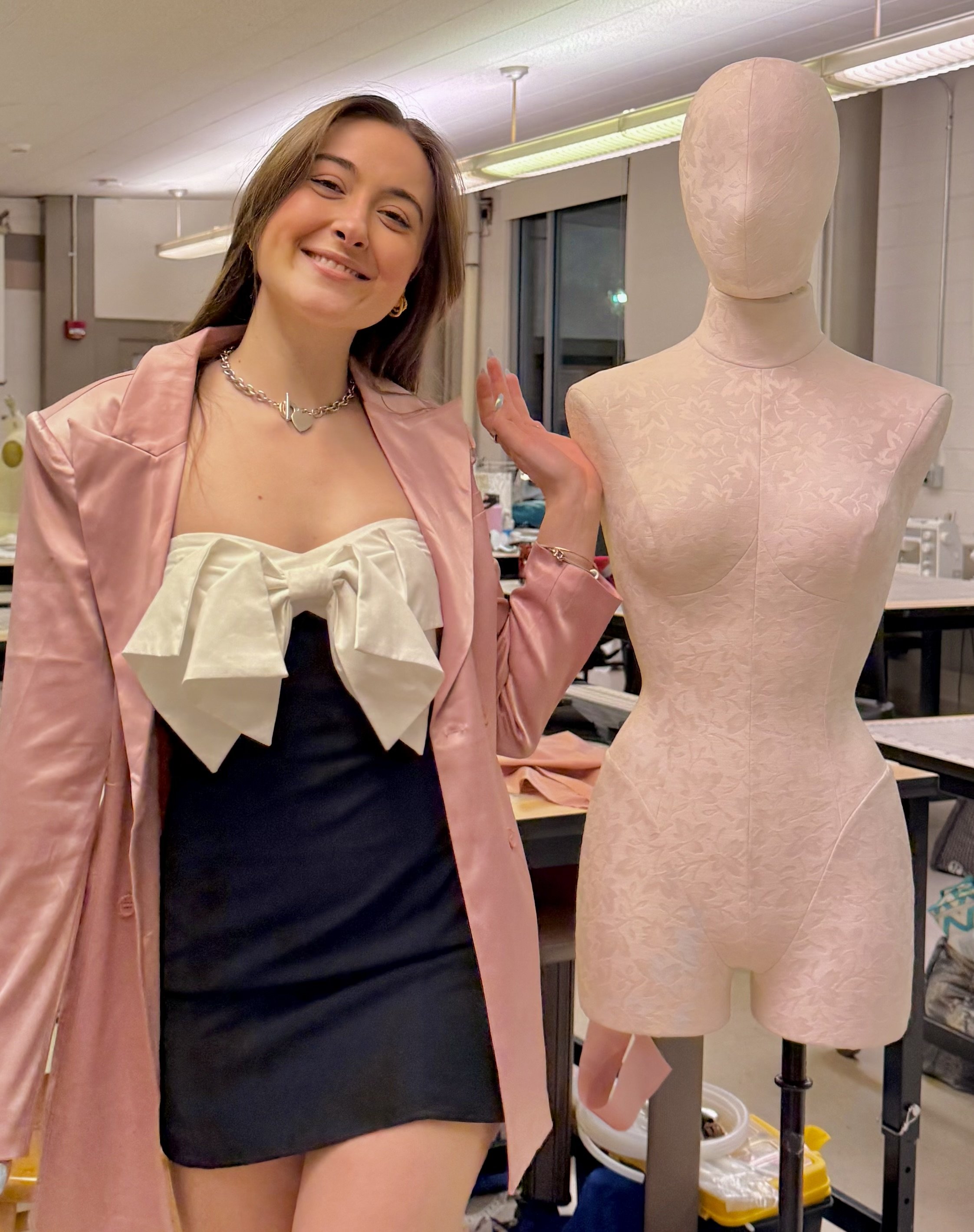 A woman dressed in a black and white dress with a pink satin jacket and necklace, smiling and standing next to a dress form with a pink fabric cover, in a room with sewing or costume workshop supplies.