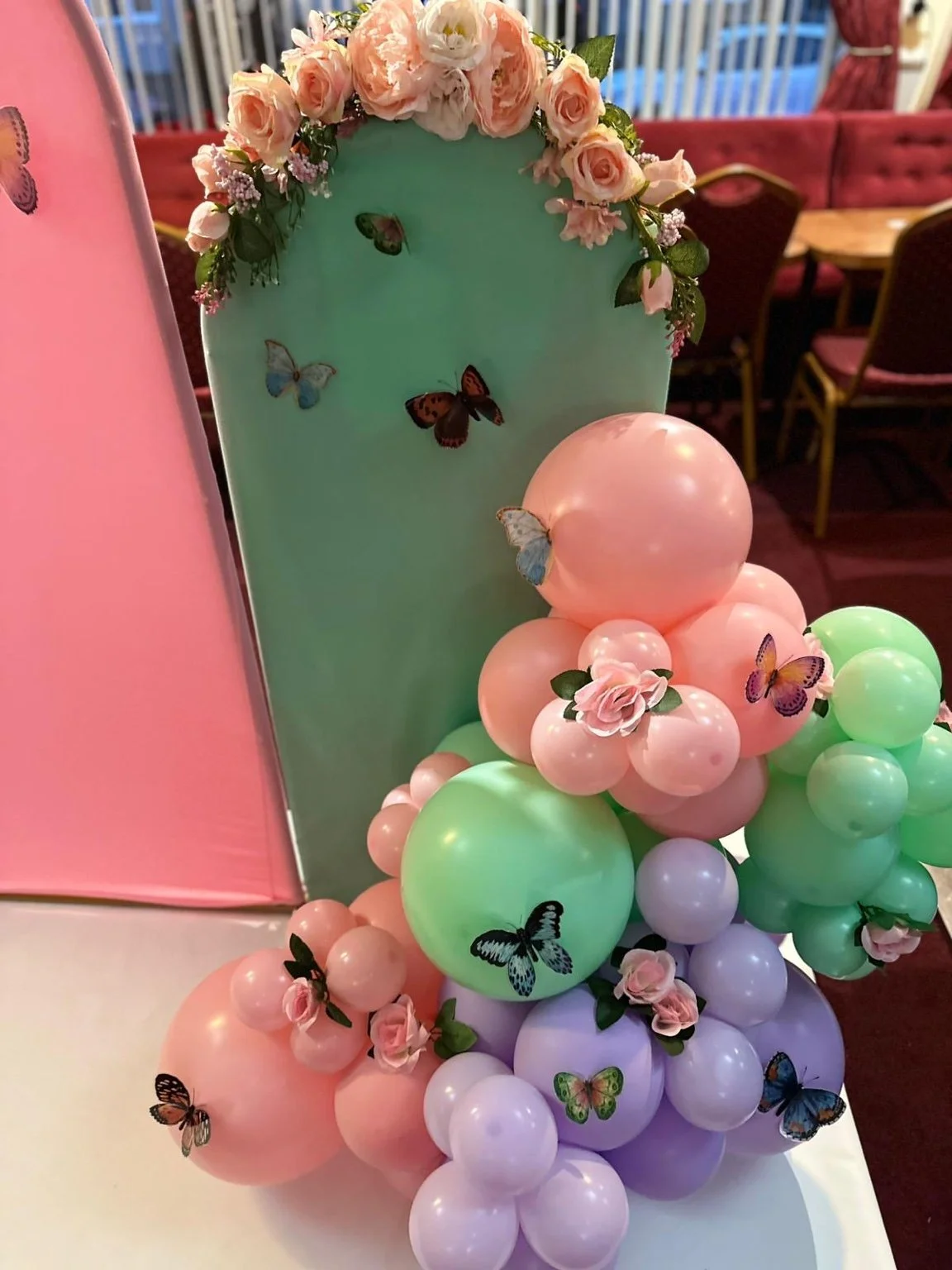Decorative balloon arrangement with pink, green, and purple balloons, adorned with artificial pink roses and butterfly stickers on a green backdrop and balloons, set against a dining area with red velvet chairs.