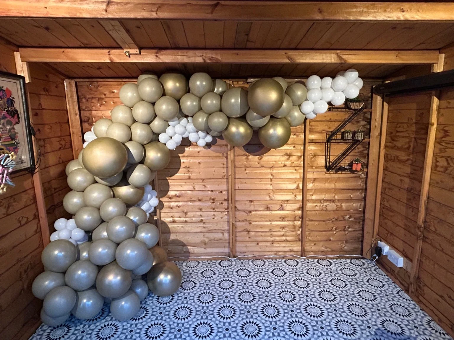 Decorative balloon arch with gold, white, and pearl-colored balloons in a wooden room with patterned black and white floor tiles.