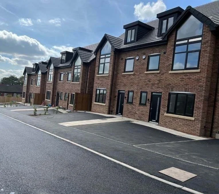 A row of newly built brick townhouses with black roofs, large windows, and small front gardens, located on a paved street.