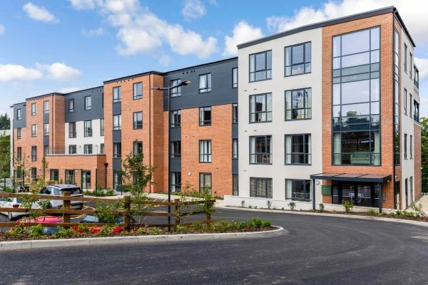 Modern multi-story apartment building with brick and white exterior, large windows, and a landscaped parking area with cars and trees.