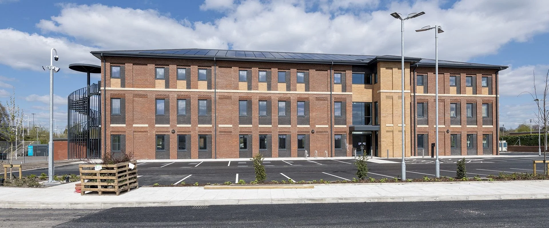 Modern four-story brick apartment building with parking lot and young trees in front, under partly cloudy sky.