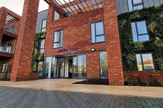 Exterior view of a modern brick apartment building named 'Ashley View' with glass entrance doors and multiple windows. Green shrubbery is visible on the side.