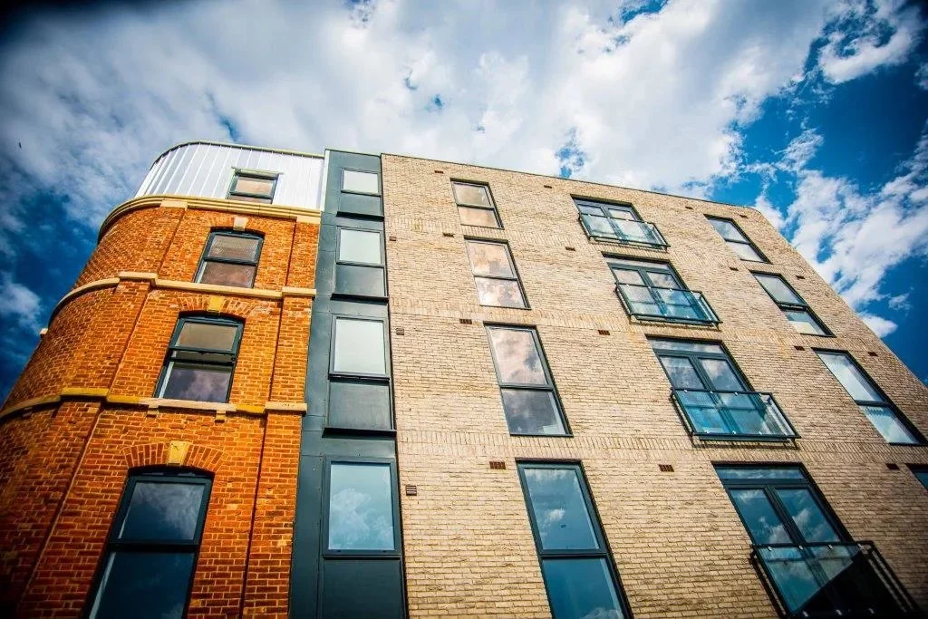 Low-angle view of a modern multi-story building with mixed brick and metal exterior, several windows, and balconies against a partly cloudy sky.