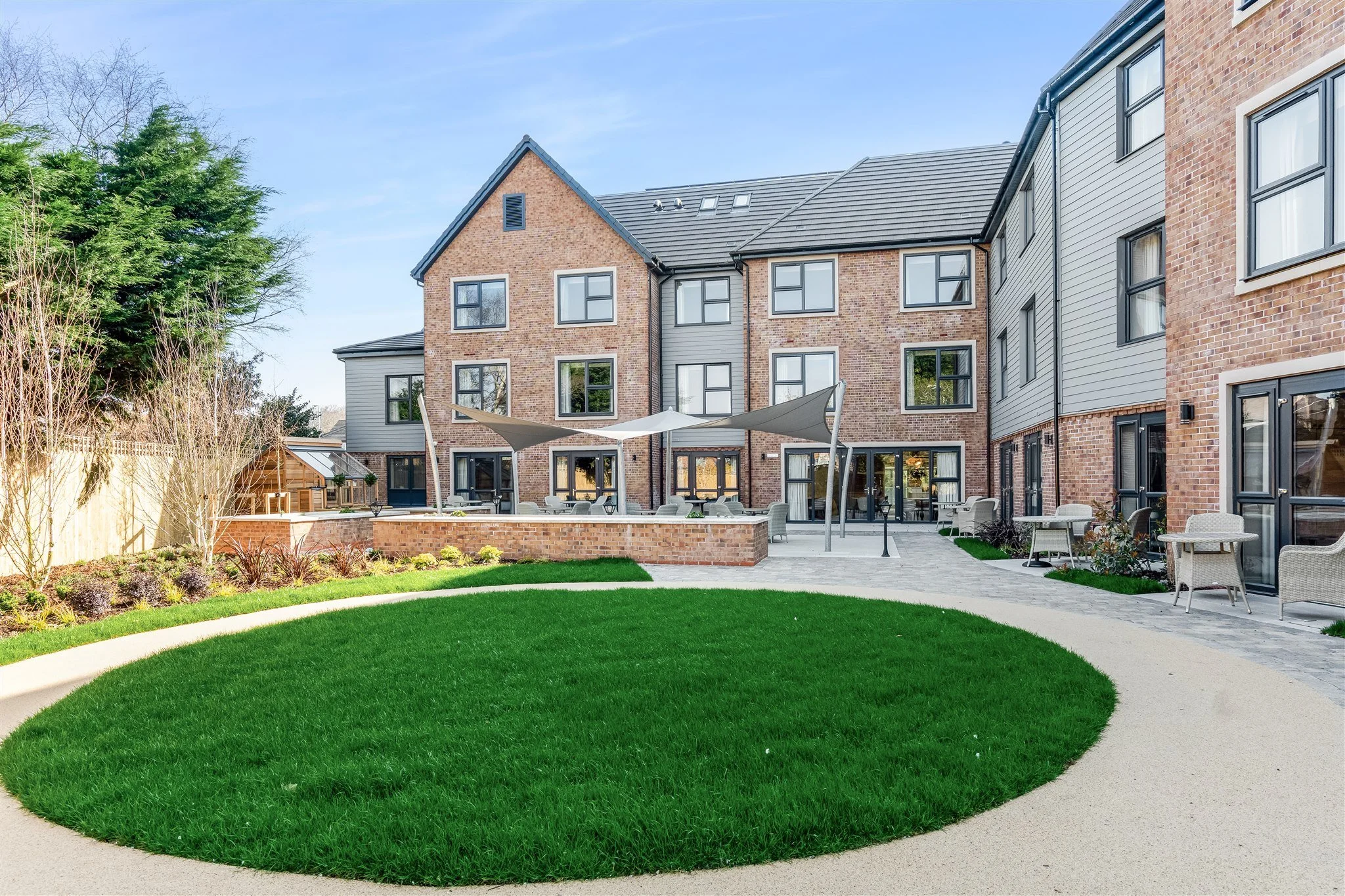 Outdoor patio area with tables, chairs, and a shaded canopy in front of a multi-story brick and siding apartment building, with a grassy circle and landscaped garden