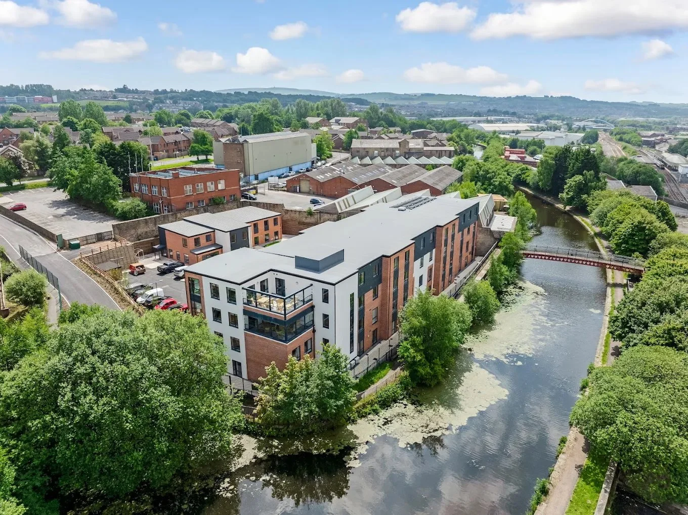 Aerial view of a small urban area with modern apartment buildings beside a river, surrounded by trees and industrial buildings, under a partly cloudy sky.