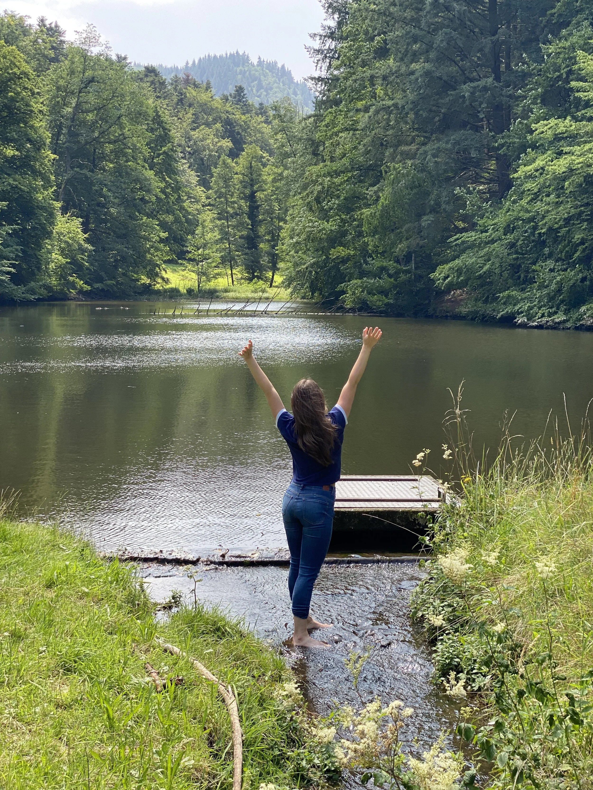 Sem T. Nova steht mit ausgestreckten Armen im Wasser am Ufer eines Sees in einem Wald.