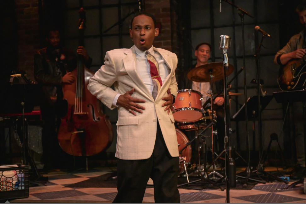 A man in a light-colored blazer with a playful expression, holding his stomach and standing in front of a band with drums and guitars on stage.