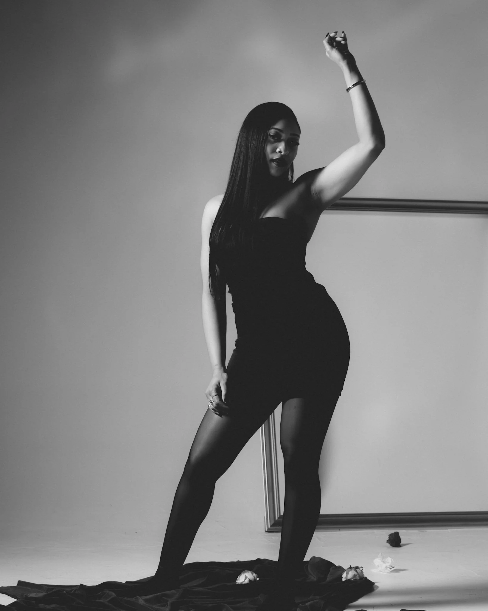 A woman with long dark hair posing in a black strapless dress, with one arm raised, standing on fabric with scattered flowers, in a photo taken against a plain background.