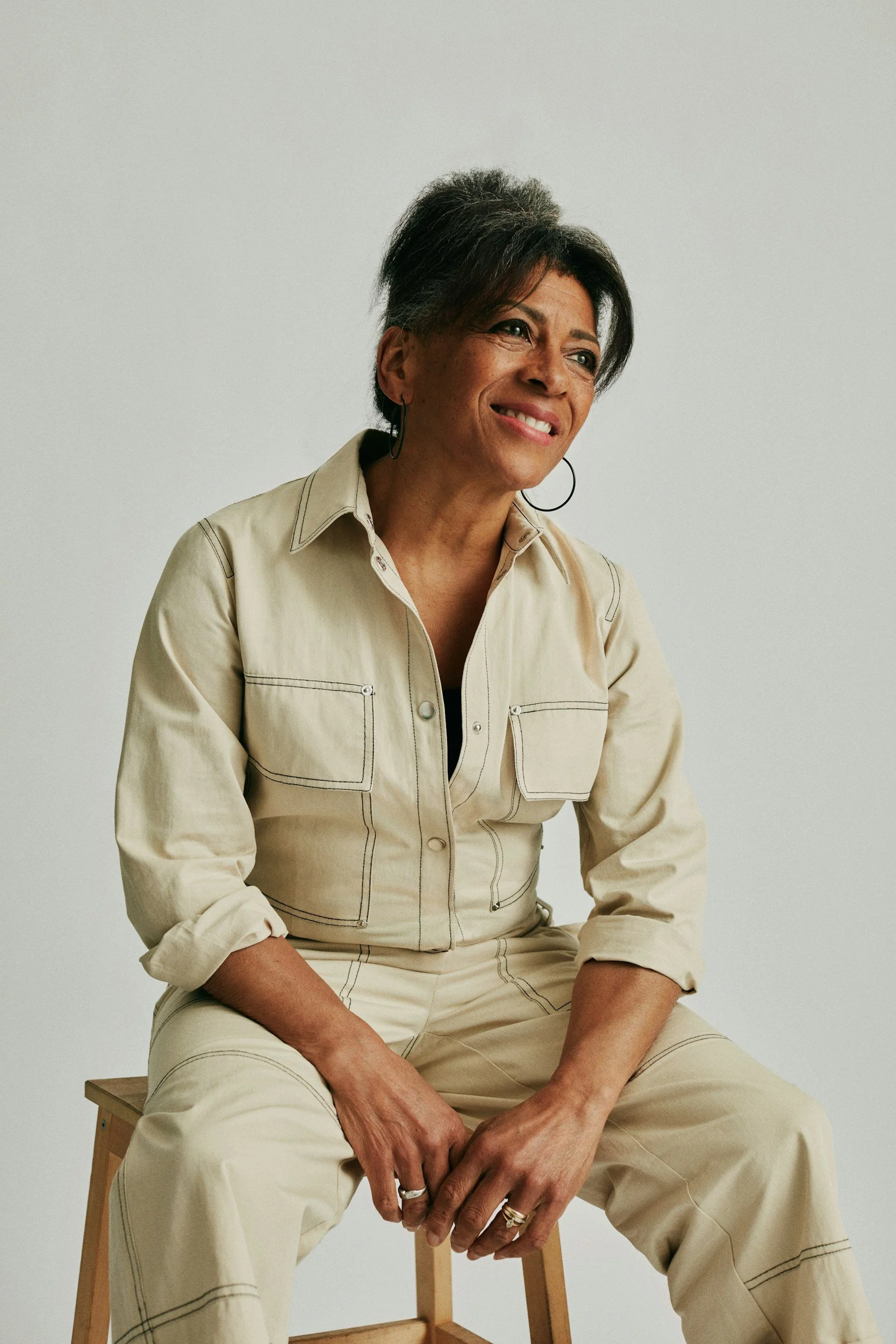 Smiling woman with short dark hair seated on a wooden chair, wearing a cream utility-style outfit and hoop earrings against a light background