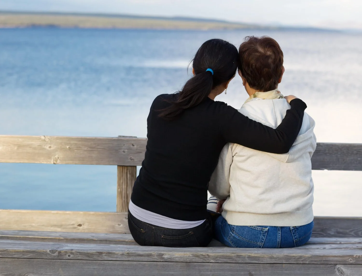 Two women sitting arm in arm on a wooden pier overlooking calm blue water