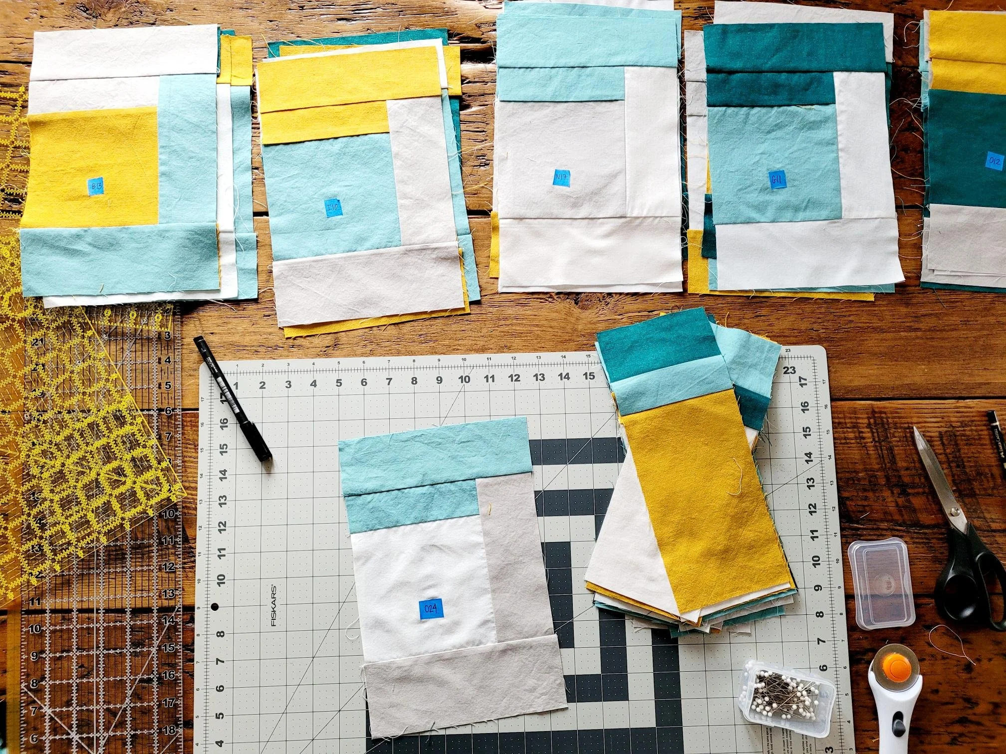 Multiple fabric quilt blocks in yellow, teal, and white colors arranged on a wooden work surface, with quilting tools including rulers, scissors, pins, a pen, and a rotary cutter.