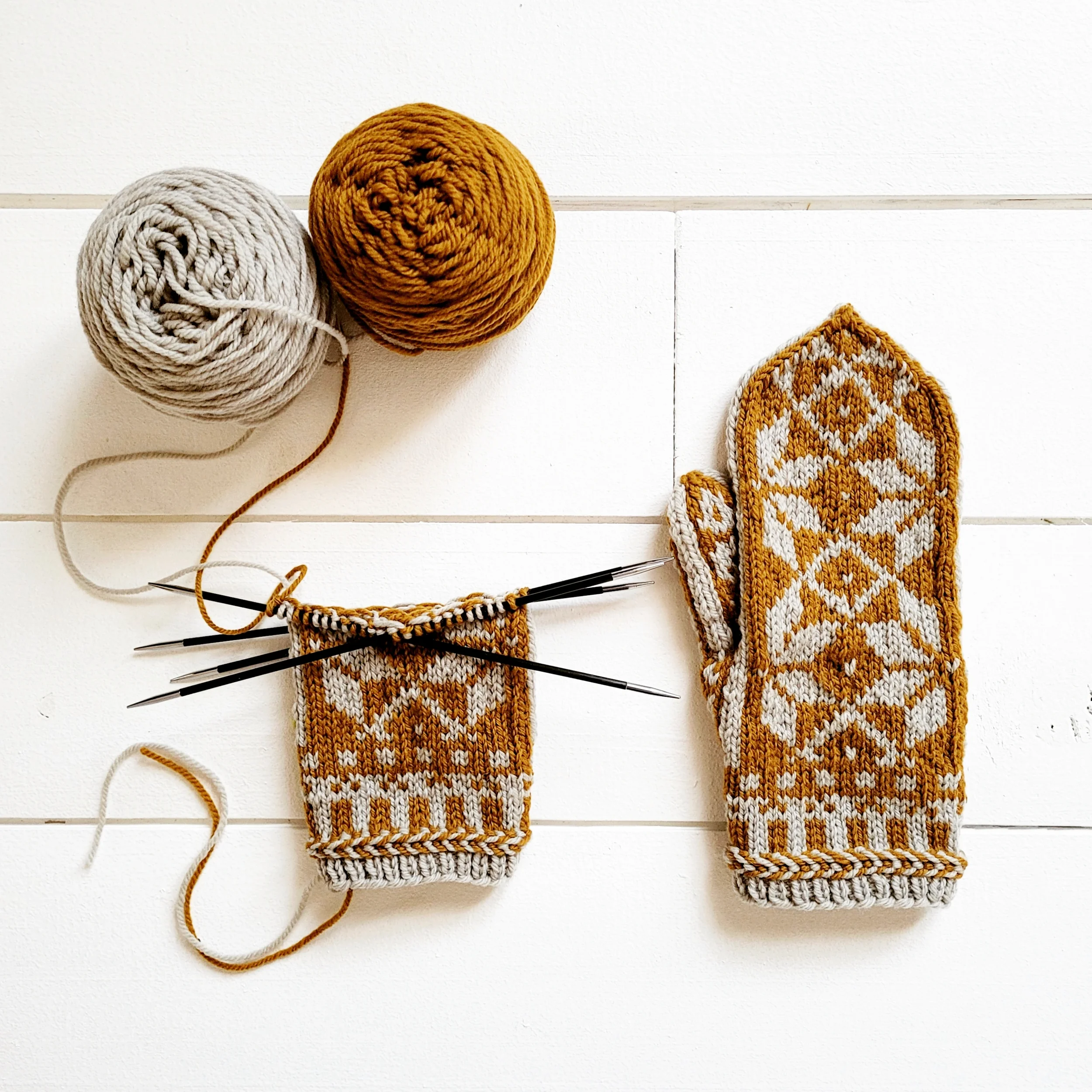 Knitted mittens in progress with yarn balls and knitting needles on a white surface.