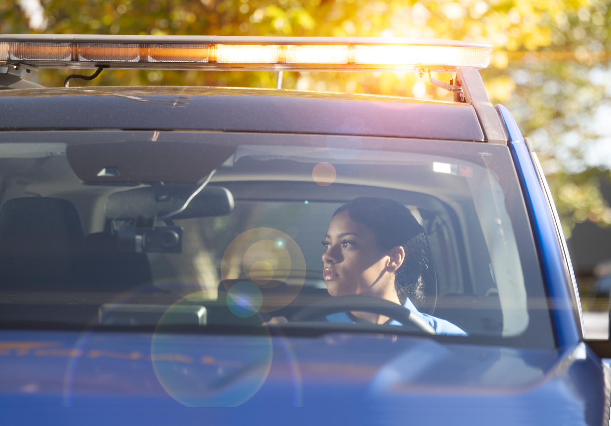 A female security officer sitting in the driver's seat of a patrol car with a light bar on top, looking to the side during daylight with trees and sunlight in the background.