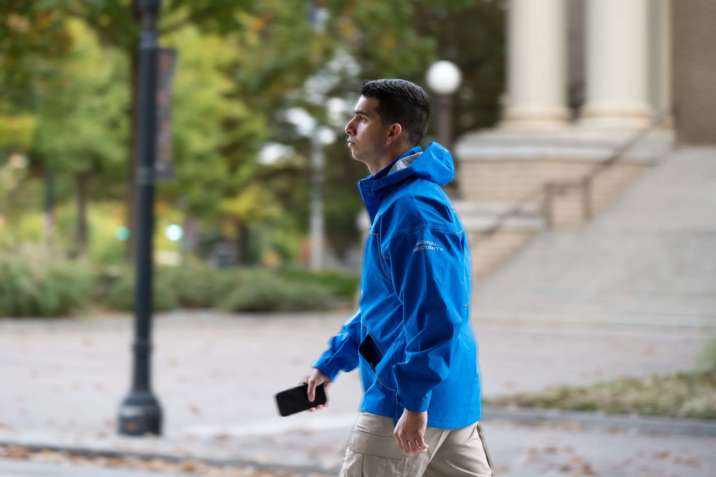 Security officer walking outdoors in a park or campus area, wearing a blue jacket labeled 'Signal Security', holding a smartphone, with trees and steps in the background.
