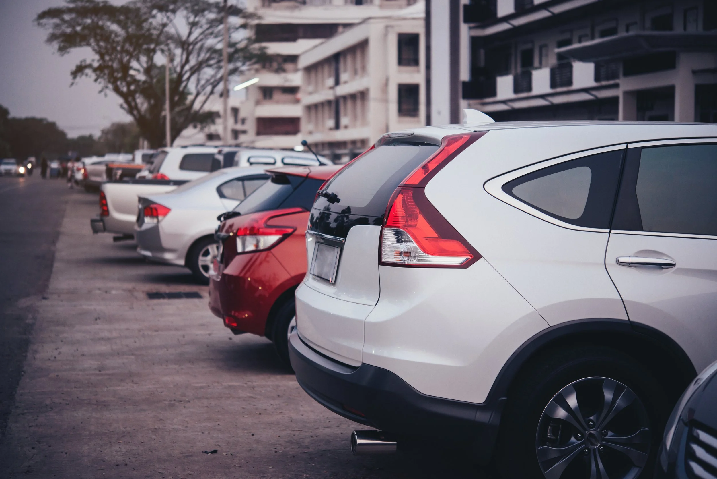 Line of parked cars along a city street with buildings and trees in the background.