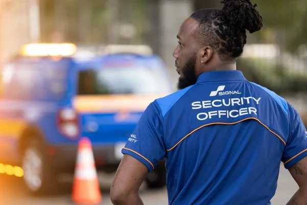 Security officer with long dreadlocked hair in a blue uniform standing outdoors near a patrol vehicle and orange traffic cones.