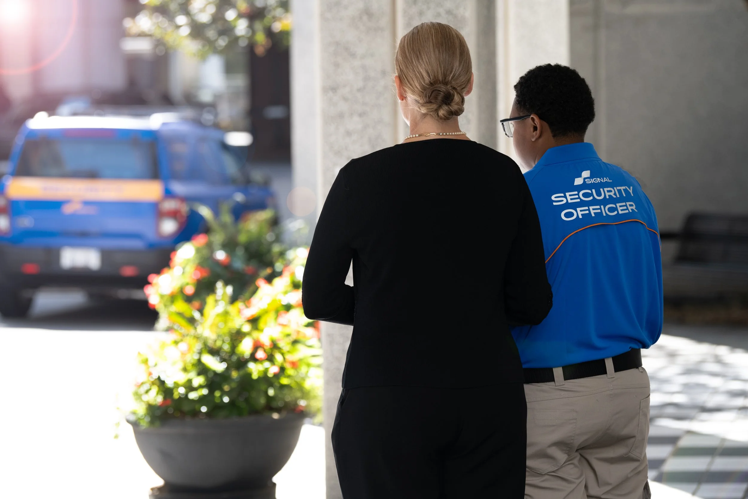 A woman in black and a security officer in blue talking outside a building, with a patrol vehicle in the background.