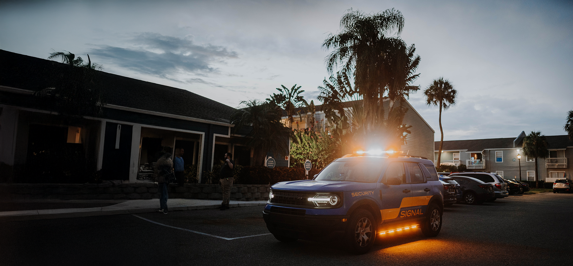 Security vehicle with flashing lights parked outside an apartment complex at dusk, with a few people standing nearby and other parked cars in the background.