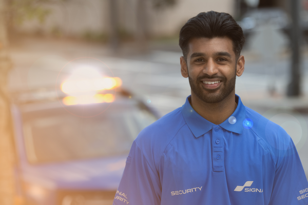 Friendly security guard in blue uniform standing outside with patrol car in the background.
