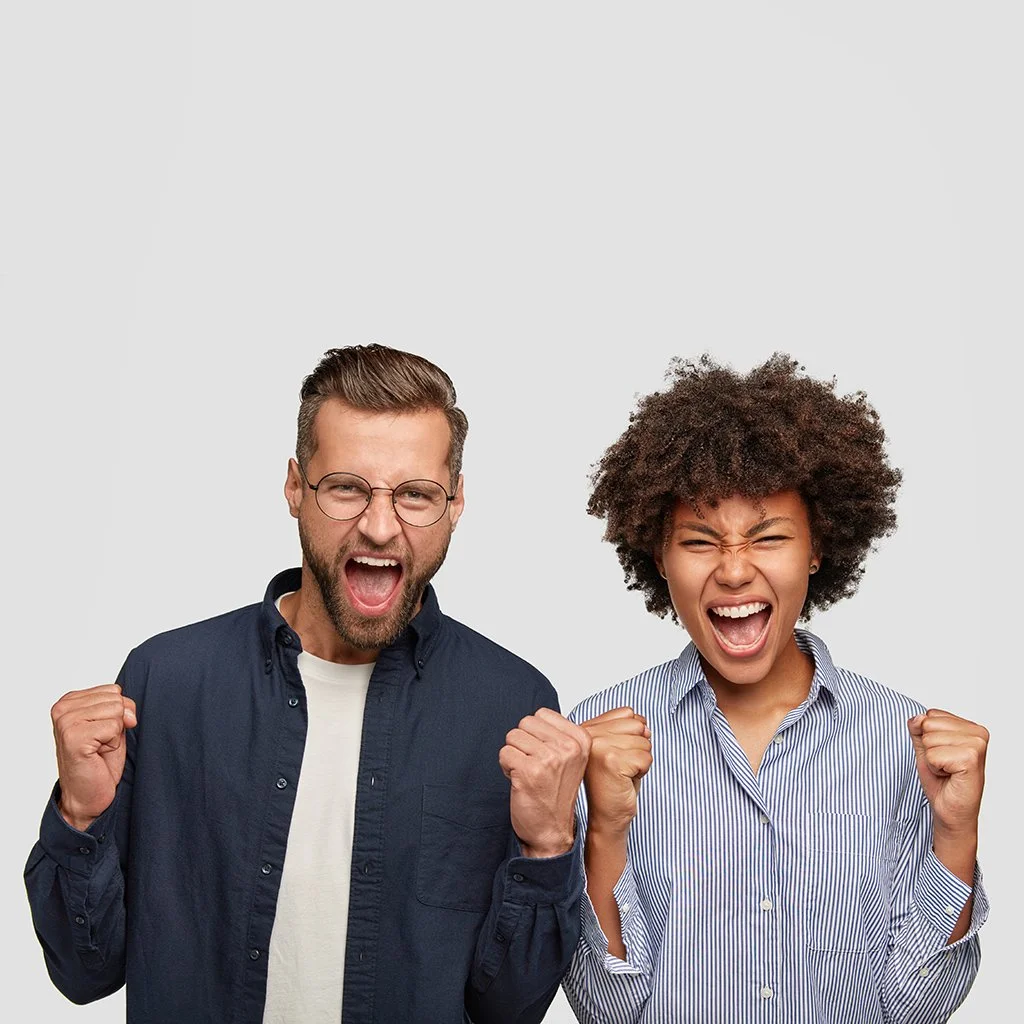 A man and woman celebrating with fists in the air and shouting excitedly against a plain light gray background.